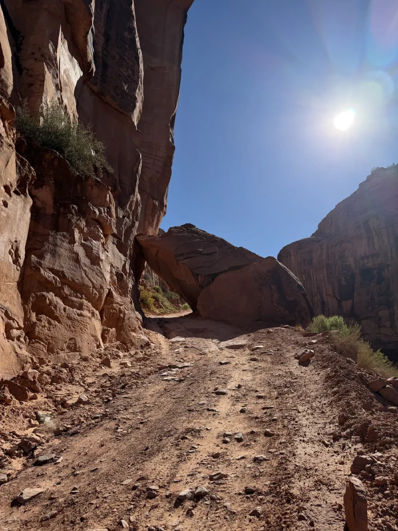 Dirt road climbing toward a huge fallen boulder wedged against a cliff wall
