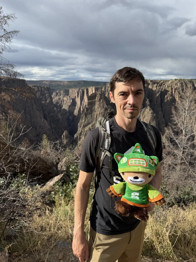 Nicholas standing on a rocky brush-covered slope below the canyon rim holding Sumi, sheer layered walls plunging behind him under heavy cloud