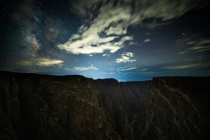 The silhouetted Black Canyon at night under long illuminated streaks of cloud with the Milky Way showing through the gaps and a cyan glow of distant town light on the horizon