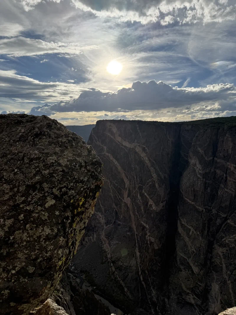 A large lichen-covered boulder perched at the cliff edge with the canyon dropping away beyond it and sun breaking through high cloud