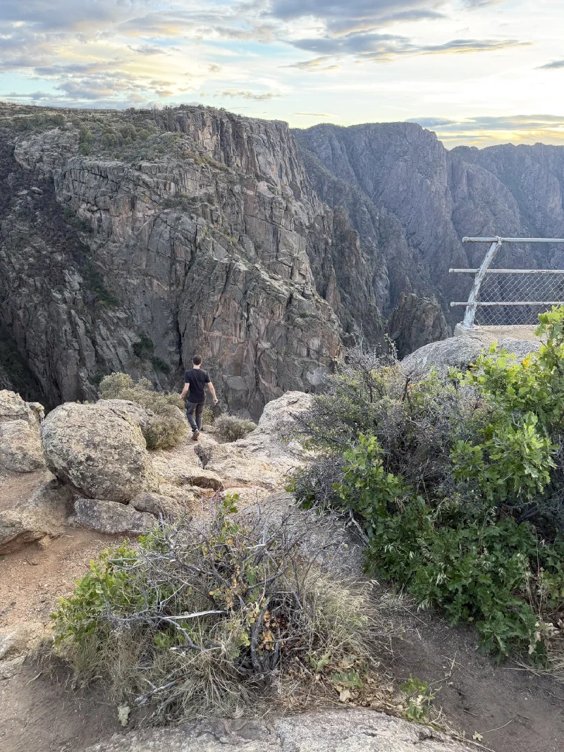 Nicholas walking down a rocky slope away from the camera toward the canyon edge, a chain barrier visible at the right