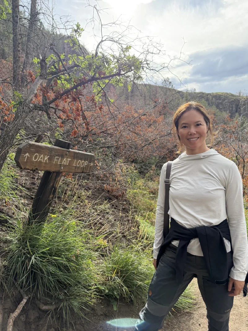 Pokin standing on a dirt trail beside a wooden sign reading OAK FLAT LOOP with an arrow, green grass and canyon rim behind