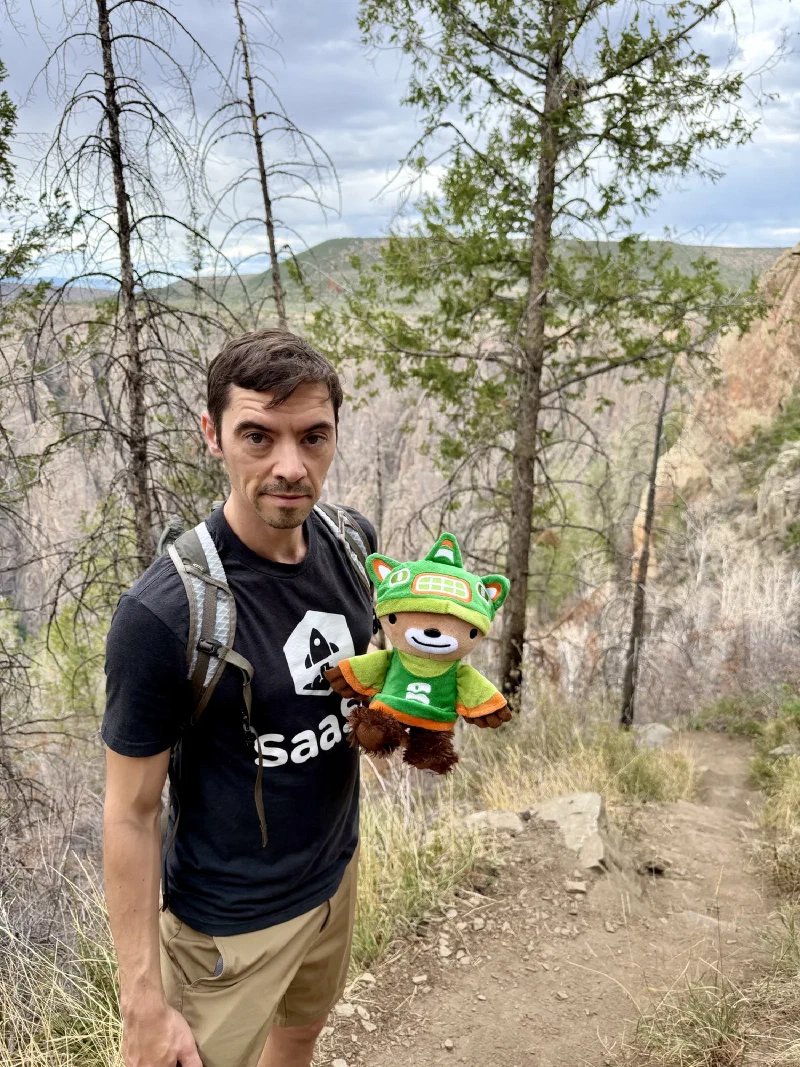 Nicholas holding Sumi on a narrow dirt trail descending between thin bare trees, canyon walls visible through the gaps