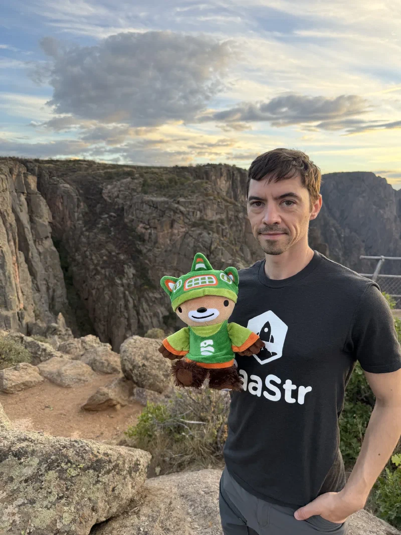 Nicholas holding Sumi at a rocky overlook with a metal railing to one side, sunset light in the textured cloud above the canyon