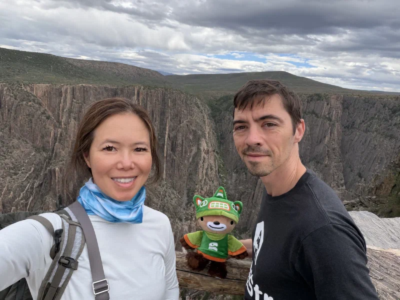Pokin taking a selfie with Nicholas, Sumi propped on the log fence between them, the canyon and cloudy sky behind