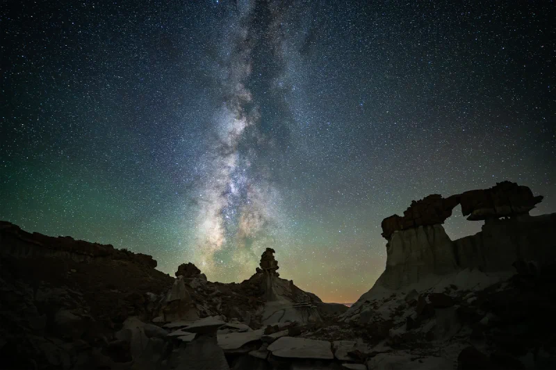 The Milky Way core standing vertically over the Bisti badlands with a large hoodoo arch on the right and pale eroded rock in the foreground