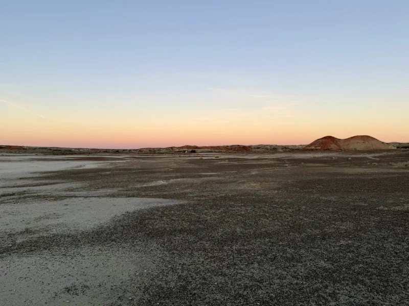 A wide flat expanse of grey gravel and pale clay stretching to low badland ridges, under a pink and orange sunset sky