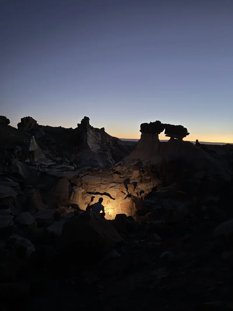 A silhouetted figure crouched among hoodoos at deep dusk with a warm headlamp glow lighting the rock around him and a rock arch on the skyline