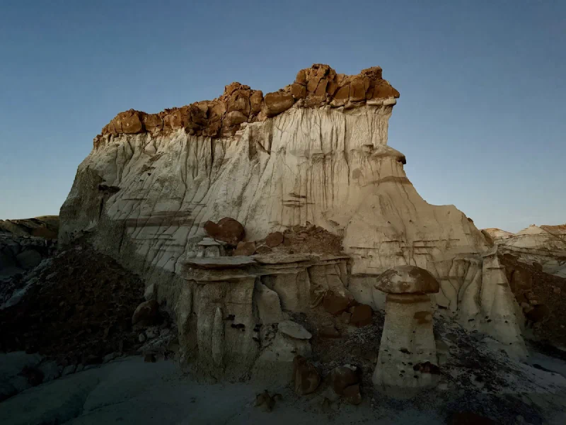 A large pale cliff with a brown boulder rim and a single mushroom shaped hoodoo standing in front of it