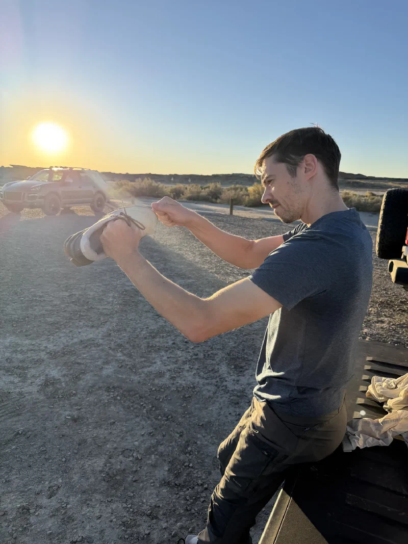Nicholas sitting on a wooden bench at a dirt car park, holding a shoe upside down and shaking sand out of it with the low sun behind him