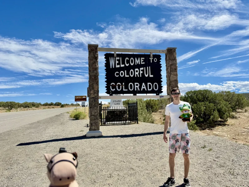 Nicholas standing beside the Welcome to Colorful Colorado state line sign holding Sumi, with Chestnut the toy horse enormous in the foreground