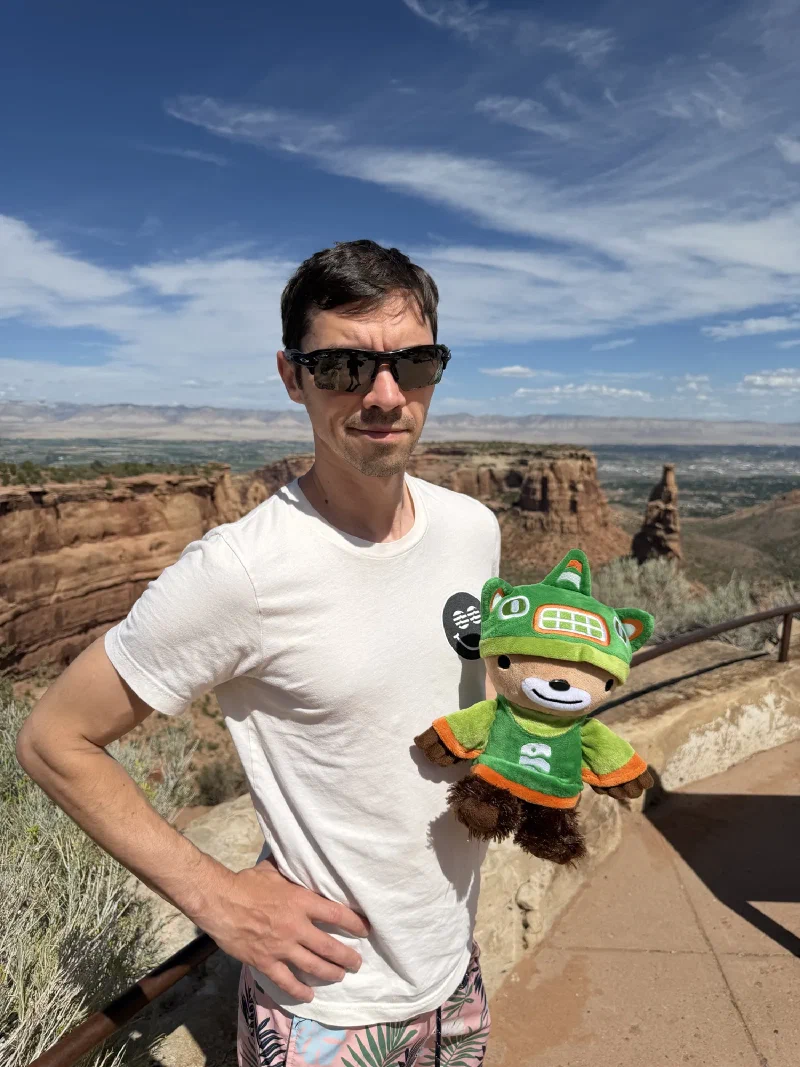 Nicholas in sunglasses at a canyon rim overlook with the freestanding Independence Monument tower rising behind him