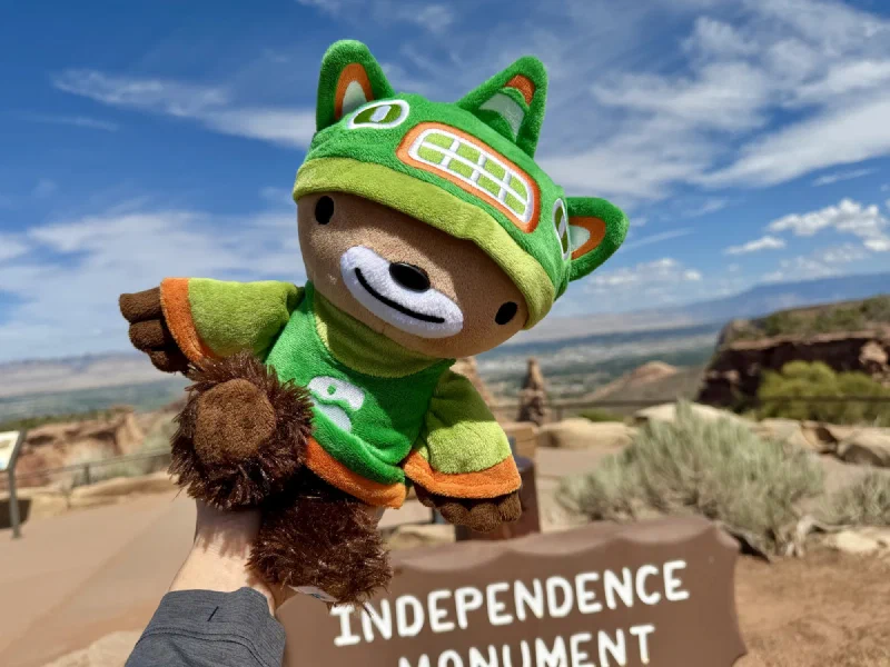Sumi held up in front of a brown park sign reading Independence Monument, with red canyon and the Grand Valley behind