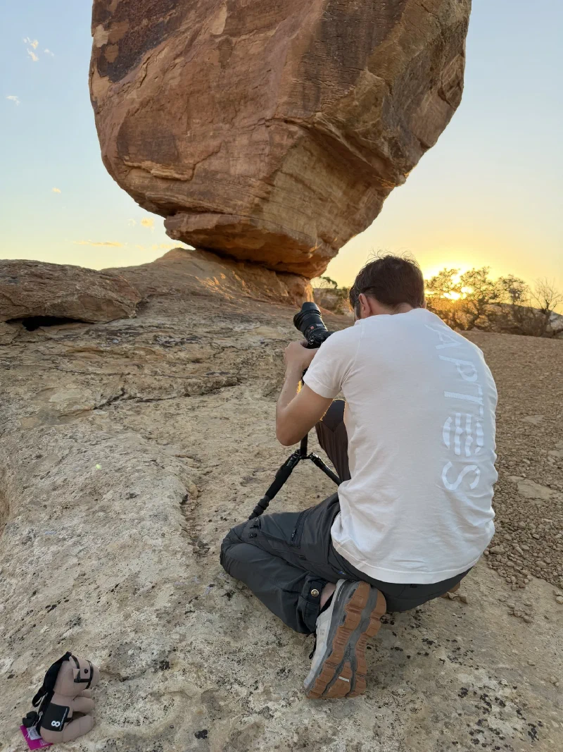 Nicholas kneeling behind his camera aimed at the balanced rock at sunset, with Chestnut the toy horse sitting on the slickrock beside him