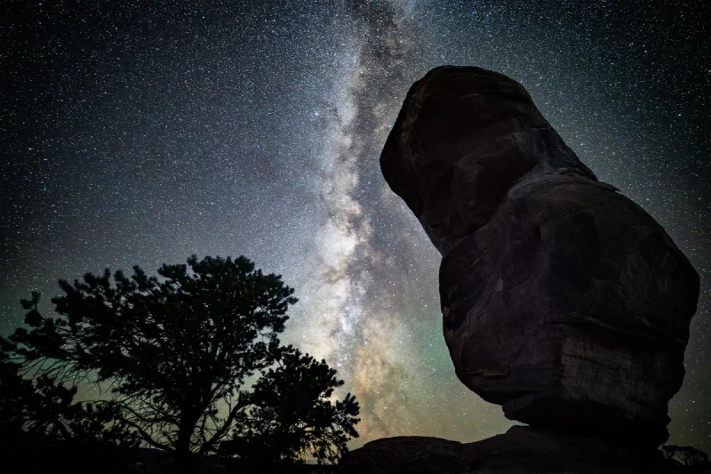 The Milky Way core rising vertically beside the silhouetted balanced rock with a juniper in the foreground