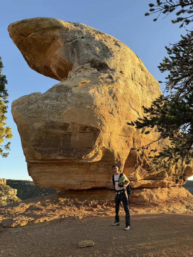 Nicholas standing at the base of the enormous balanced boulder of Miracle Rock in golden evening light