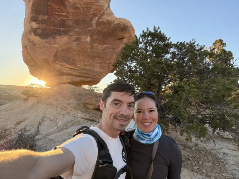 Nicholas and Pokin taking a selfie under the balanced rock with the setting sun bursting in a starburst beneath it