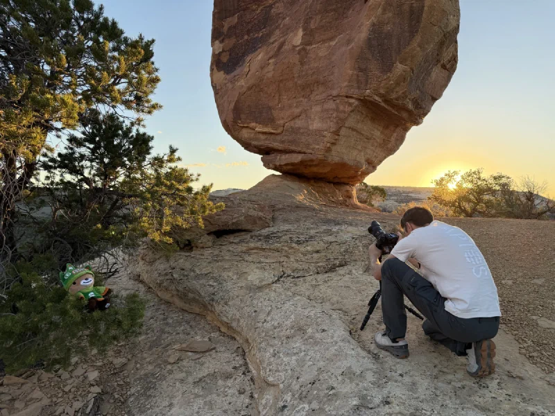A camera on a tripod below the balanced rock as the sun sets behind, with Sumi propped in a juniper bush
