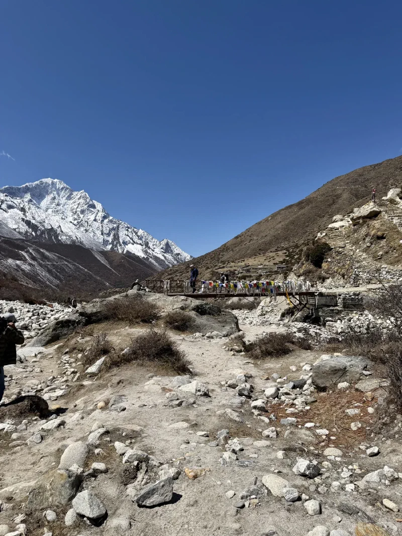 Metal bridge covered in prayer flags spanning a rocky riverbed
