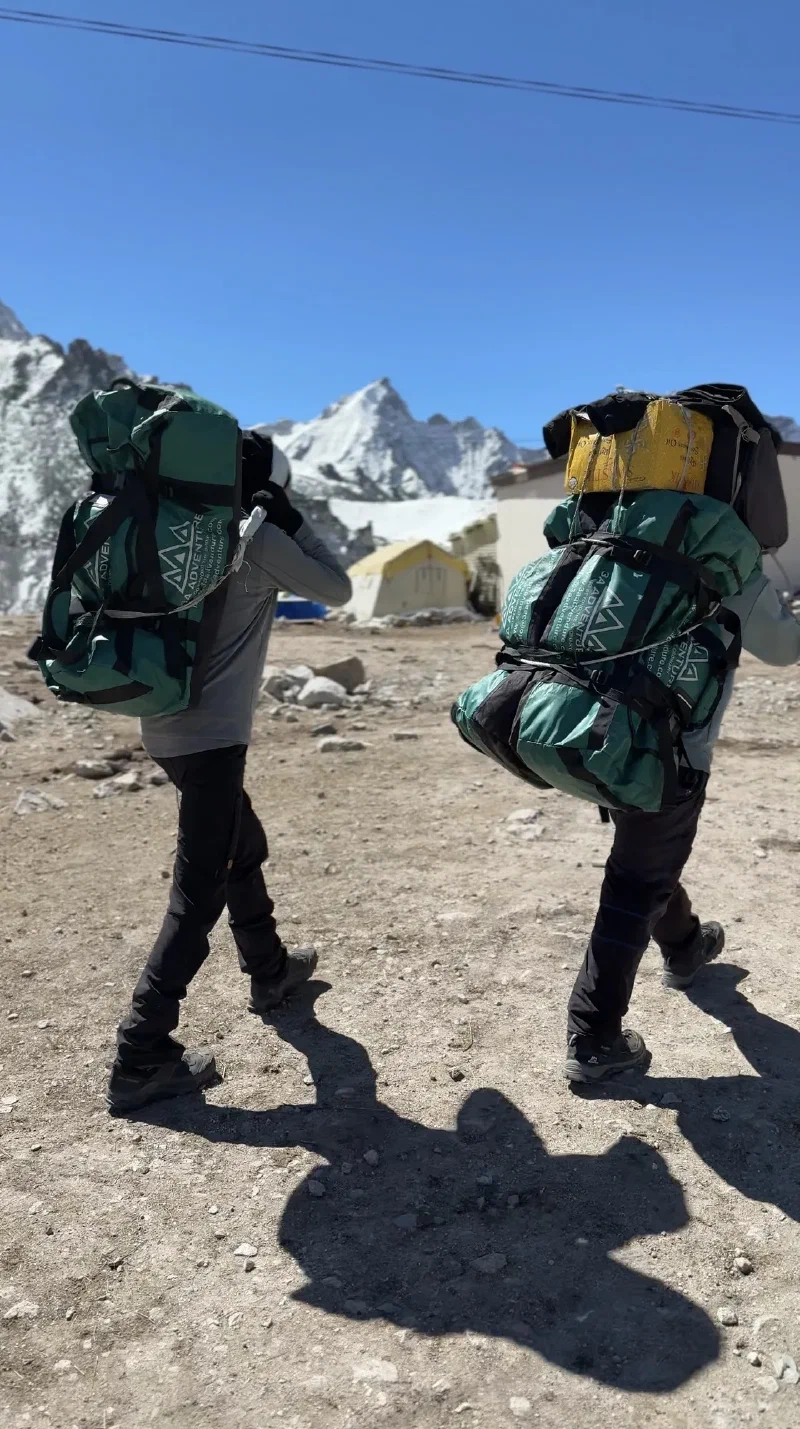 Nicholas and a porter walking away on the trail with large duffel bags and namlo straps, mountains behind