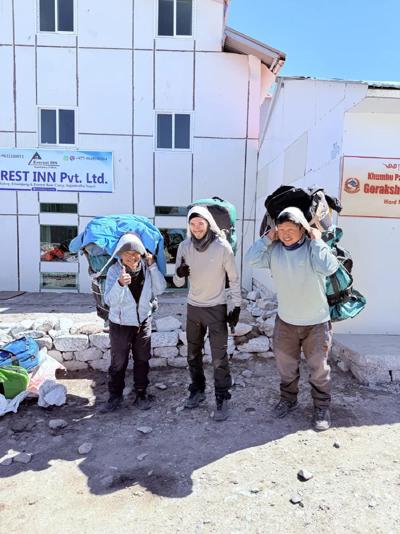 Nicholas standing between two porters outside a lodge, all carrying bags with namlo head straps