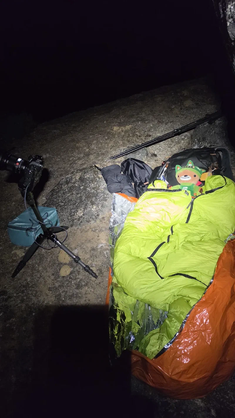 Camera on tripod next to sleeping bag and emergency blankets on a rocky hillside at night