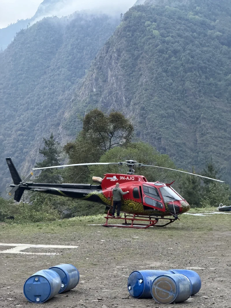A Fishtail Air helicopter on a foggy dirt helipad, pilot leaning into the cabin, blue barrels in the foreground
