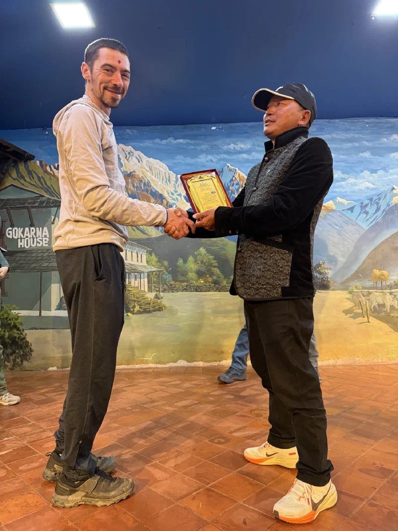 D.B. presenting Nicholas with a framed plaque on the stage, both with red tika marks on their foreheads