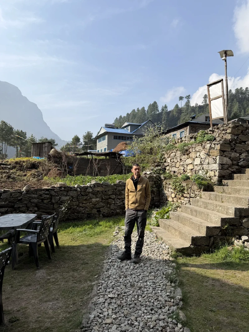 Nicholas standing in a stone courtyard with outdoor dining tables, green hillside and a mountain peak behind