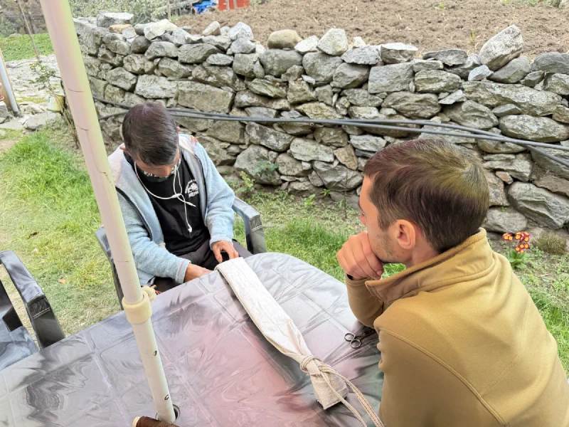 Porter Kerman sewing a head strap from a cut rice bag at an outdoor table while Nicholas watches