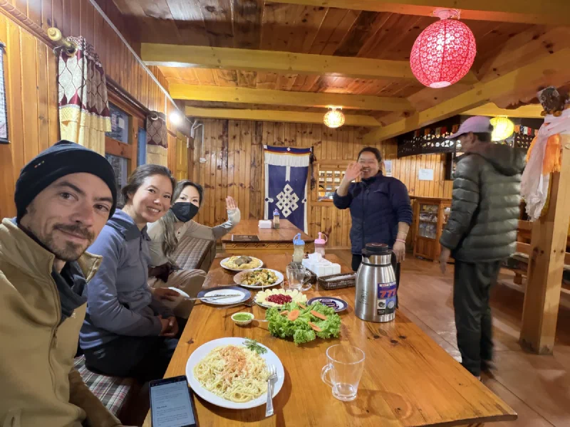 Nima Sherpa serving a big spread of food to the smiling group at a long wooden table under red lanterns
