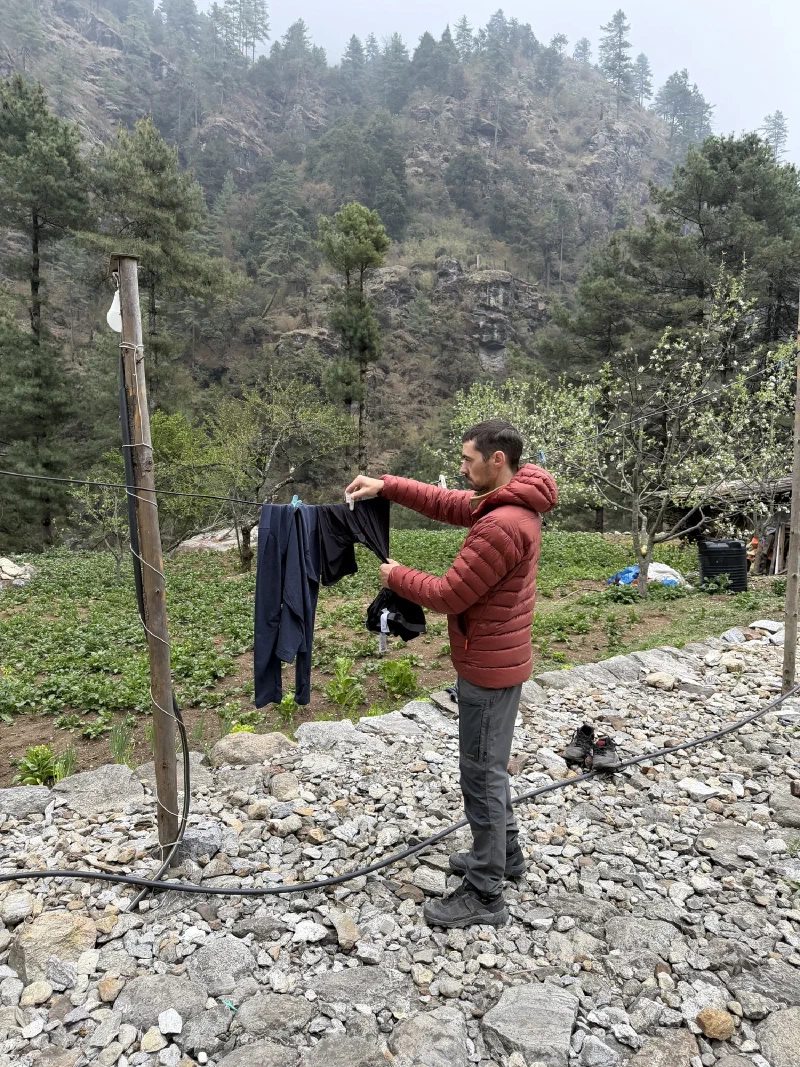 Nicholas hanging dark laundry on a clothesline with a misty pine-covered hillside behind