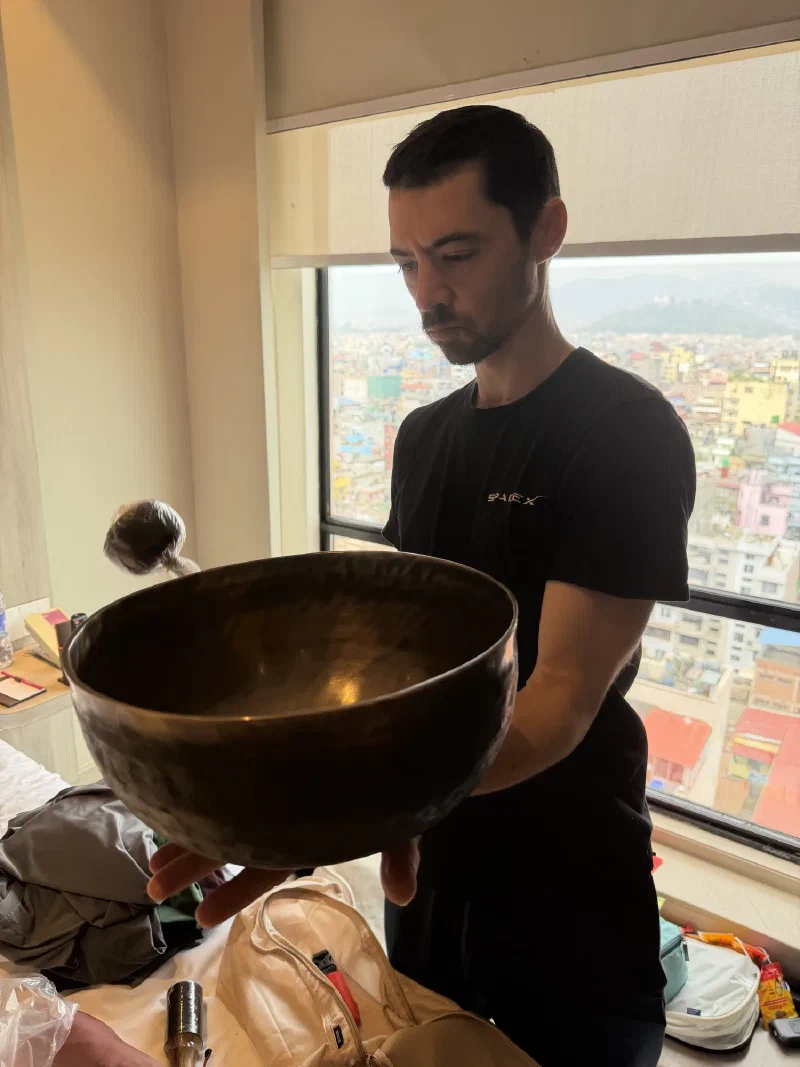 Nicholas in a SpaceX t-shirt testing a large metal singing bowl by a hotel window overlooking Kathmandu
