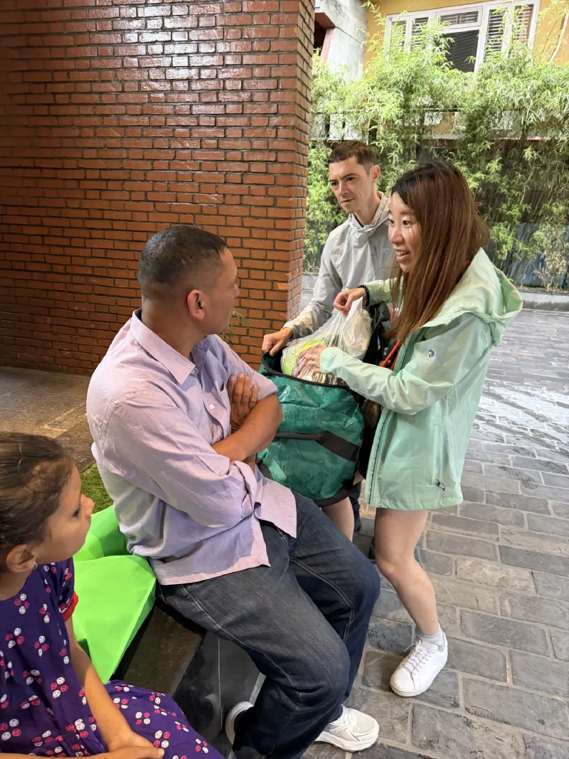 Alice unloading a plastic-wrapped package from a green duffel bag for Manoj, with Manoj's daughter watching
