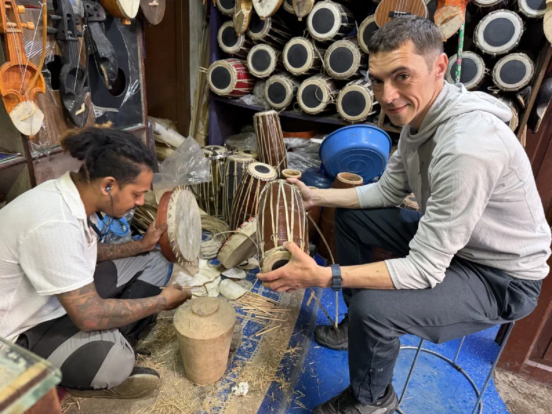 Nicholas holding a hand drum with Ashish, a tattooed drum-maker, working on a frame drum on the floor surrounded by wood shavings