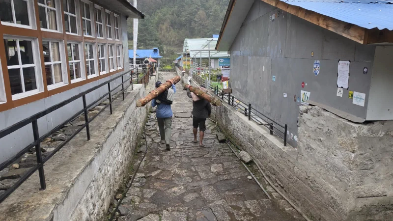Two men carrying large raw logs on their shoulders through a narrow stone village alley