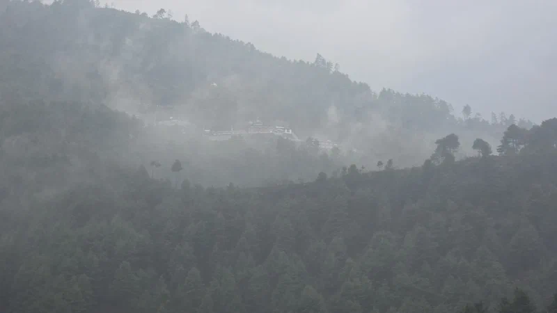 A large monastery complex visible through mist on a steep forested hillside with no obvious path