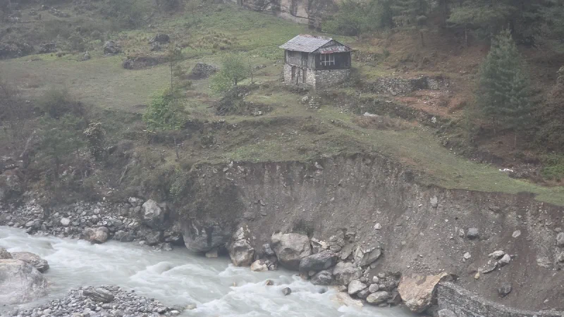 A solitary stone and wood cabin perched on a steep riverbank above a rushing glacial river