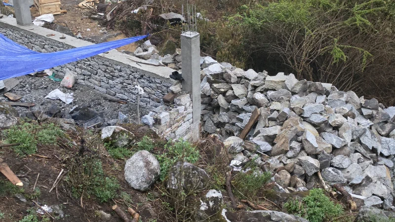 A construction site with partially built stone walls, concrete columns with exposed rebar, and piles of broken quarried stone