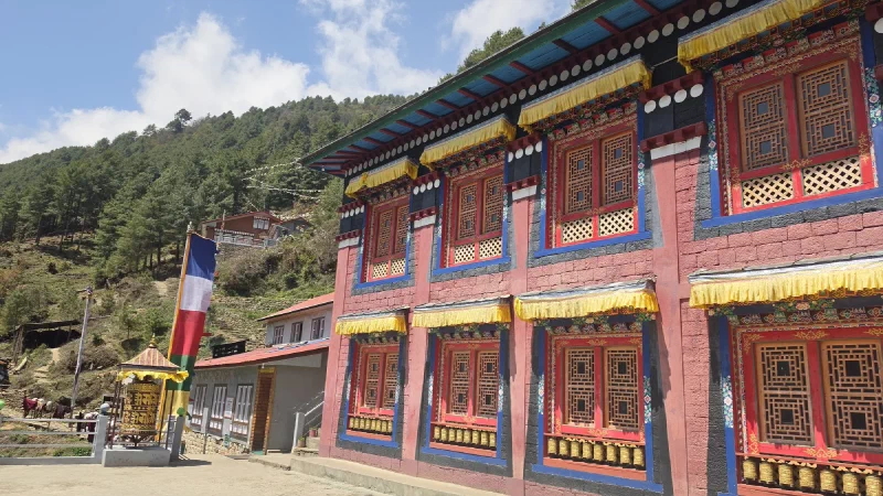 Ornate monastery building with red walls, gold trim, blue roof and a prayer wheel out front