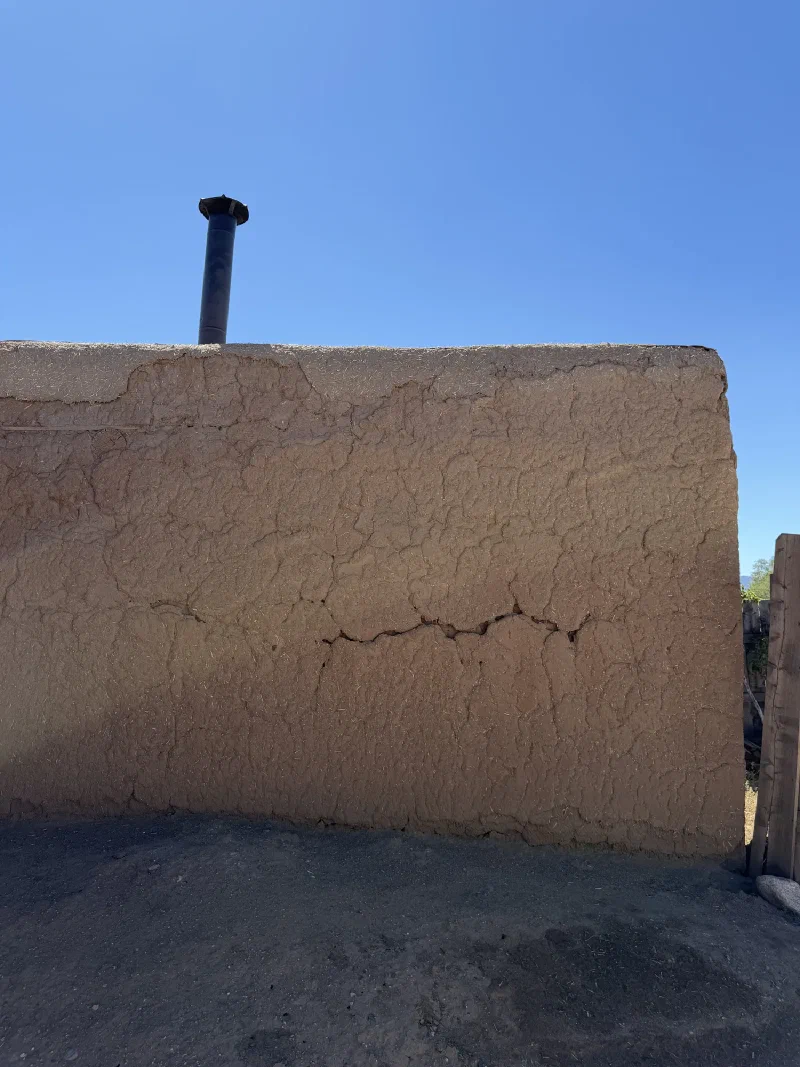 A close up of a cracked and uneven adobe wall showing straw mixed into the earth plaster, with a black metal chimney pipe protruding from the flat roof above against a clear blue sky