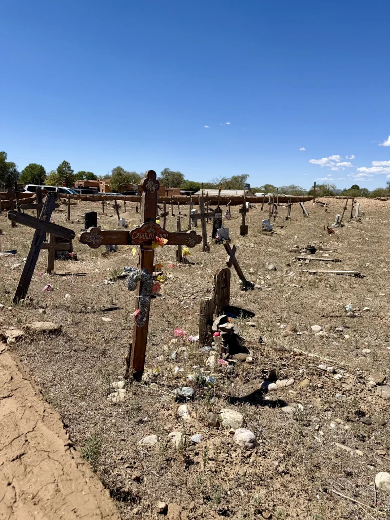 A cemetery at Taos Pueblo with many weathered wooden crosses at various angles on dry rocky ground, bordered by a low adobe wall with trees and rooftops beyond