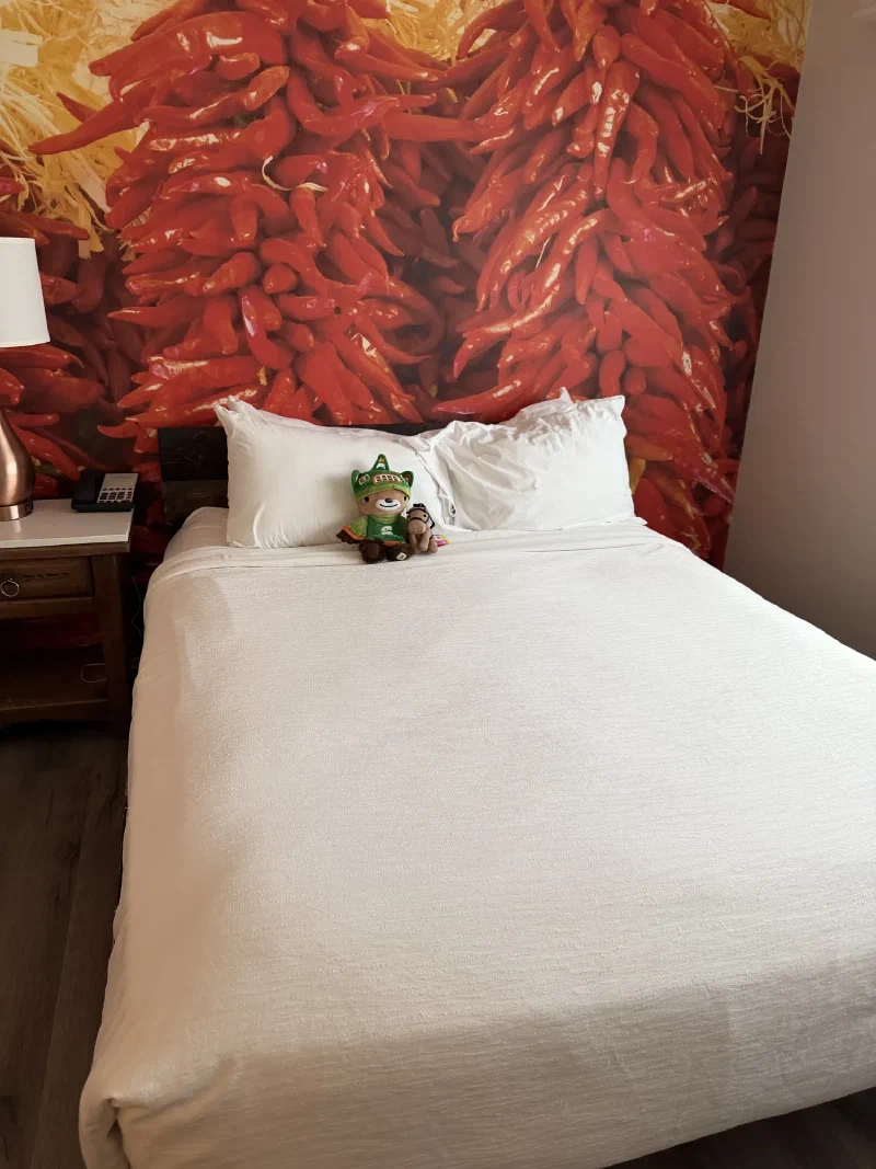 Sumi and Chestnut the toy horse propped against white pillows on a hotel bed, with a large mural of hanging red chilli ristras covering the headboard wall