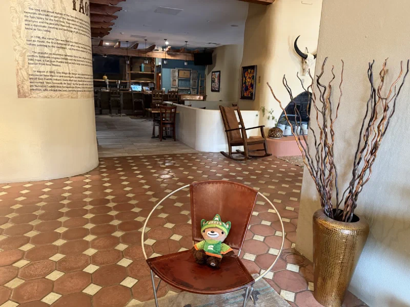 Sumi the small brown bear sitting in a round brown leather chair in a southwestern hotel lobby, with a curved wall of printed Taos history text, hexagonal tile floor, and a white adobe fireplace behind