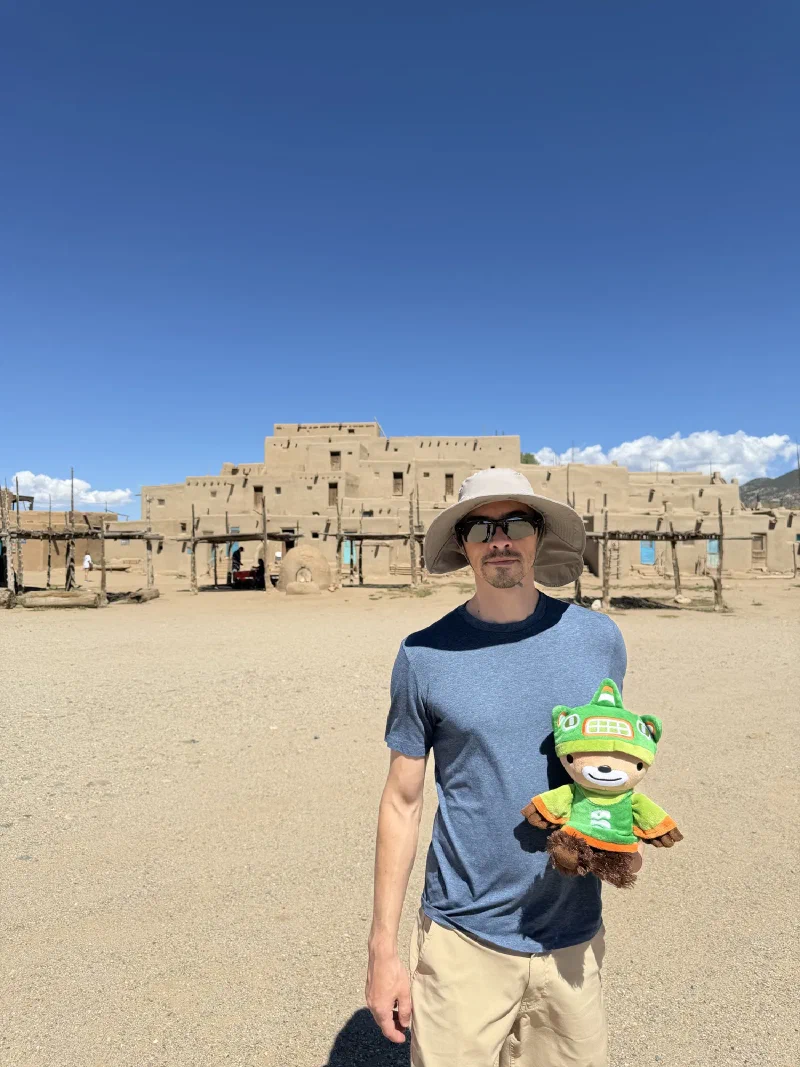 Nicholas in a grey sun hat and sunglasses holding Sumi, with the multi storey terraced adobe buildings of Taos Pueblo and their wooden ladders rising behind him