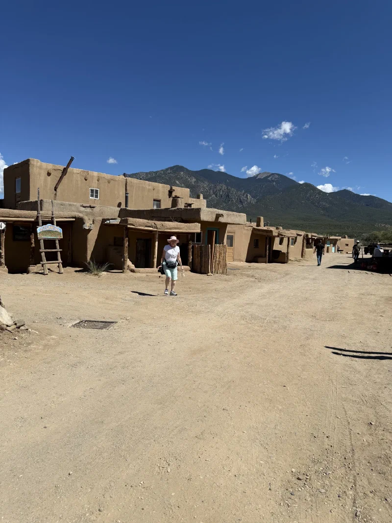 A wide dirt plaza at Taos Pueblo bordered by single storey adobe buildings with wooden porch posts, a leaning wooden ladder, a hanging sign reading Aspenwind Native Arts, and green mountains behind under a deep blue sky