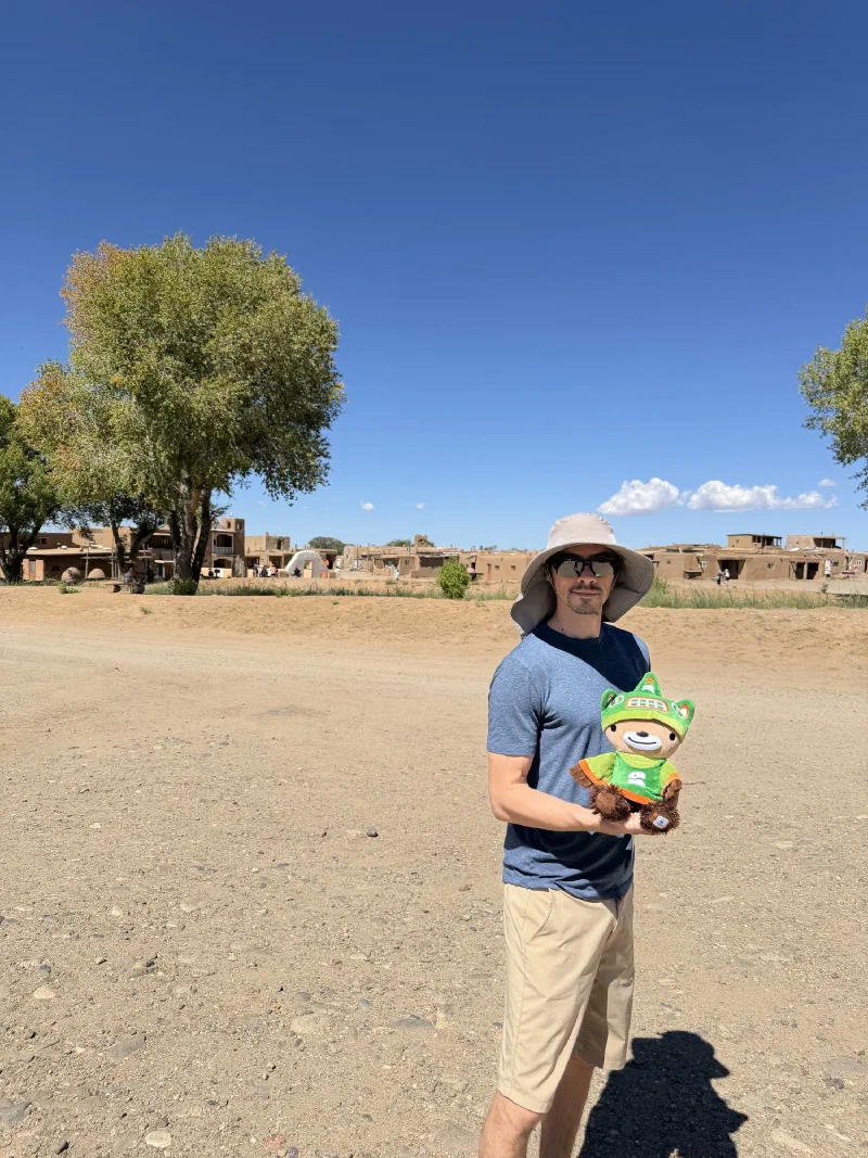Nicholas in a blue t-shirt and grey sun hat holding Sumi in an open dusty area, with a row of single storey adobe buildings and green trees across the field behind them