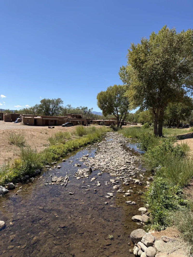 Red Willow Creek running clear over a rocky bed through Taos Pueblo, with green trees along the banks and multi level adobe buildings visible behind