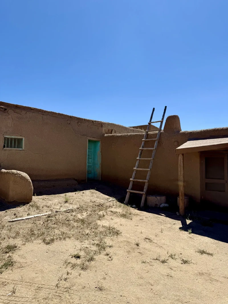 A long single storey adobe building with a turquoise painted door, a weathered wooden ladder leaning against the wall, and a rounded adobe horno bread oven in the foreground