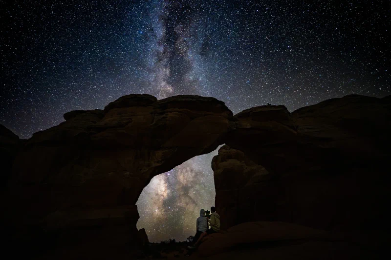 Pokin and Nicholas sitting together under Broken Arch at night holding Sumi, with the Milky Way blazing above and through the opening